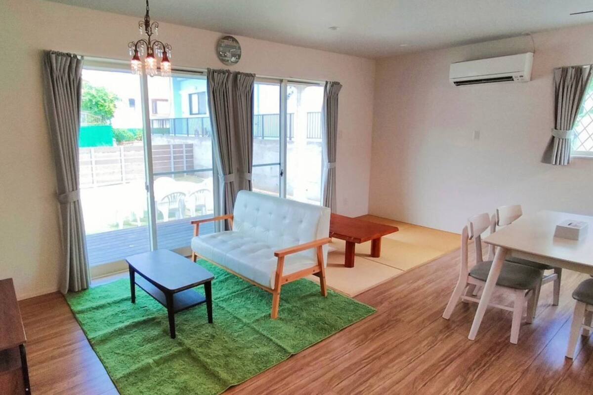 Uma sala de estar com sofá, uma mesa pequena, uma mesa de jantar com cadeiras, uma televisão sobre um móvel e uma janela grande com cortinas.