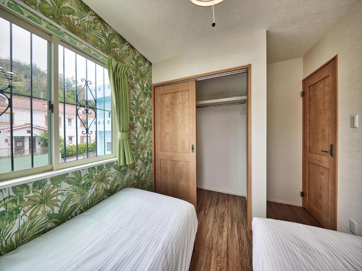 A room with two beds, a wooden wardrobe, and a window with a view of buildings.