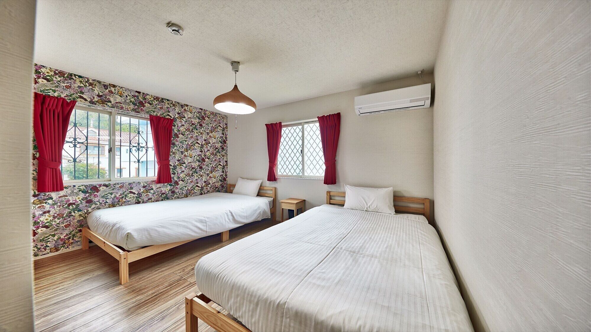 Ein Zimmer mit zwei Betten, einem kleinen Tisch, einer Hängelampe und einer Klimaanlage.
