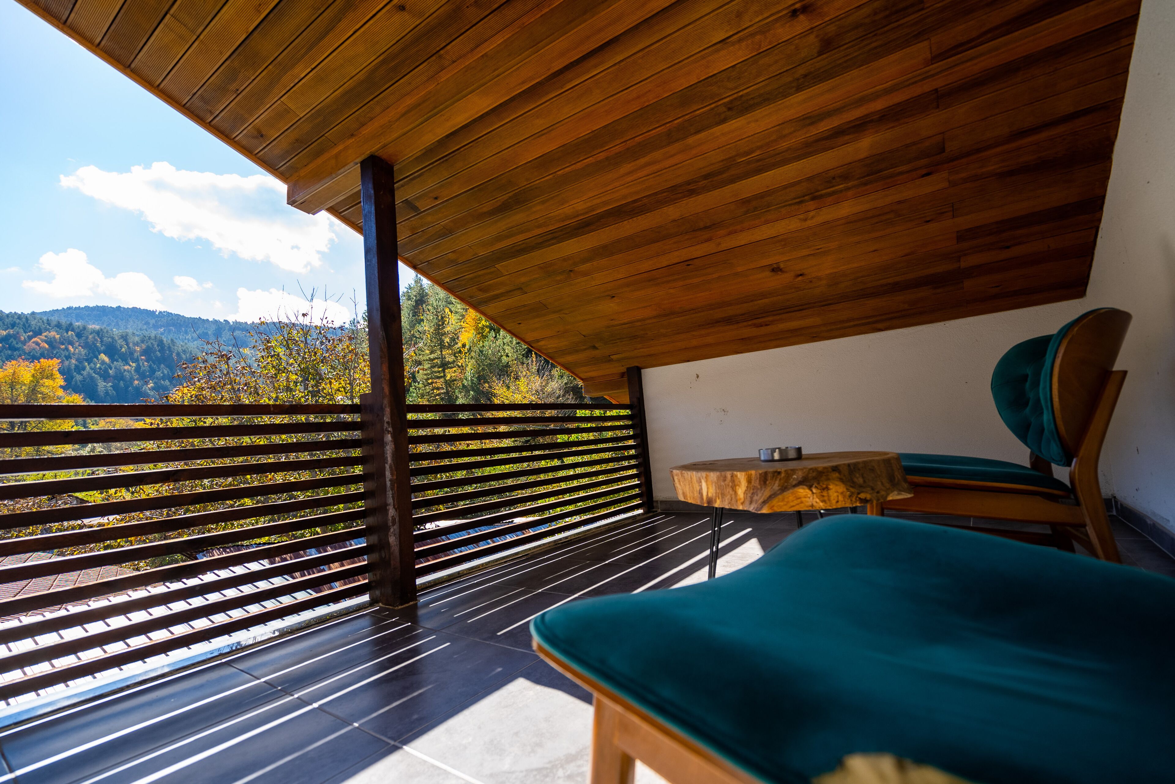 A wooden deck with a view of autumn trees, a wooden table, and a teal chair.