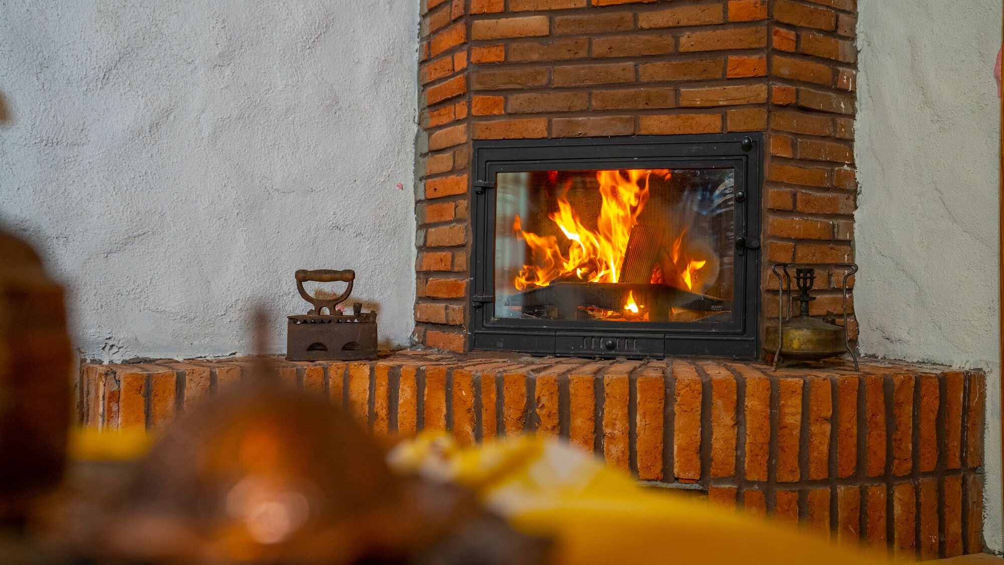 Ein Kamin aus Ziegelsteinen mit sichtbarem Feuer, ein Metall-Wasserkocher und eine kleine Dampfbügeleisen auf dem Sims.