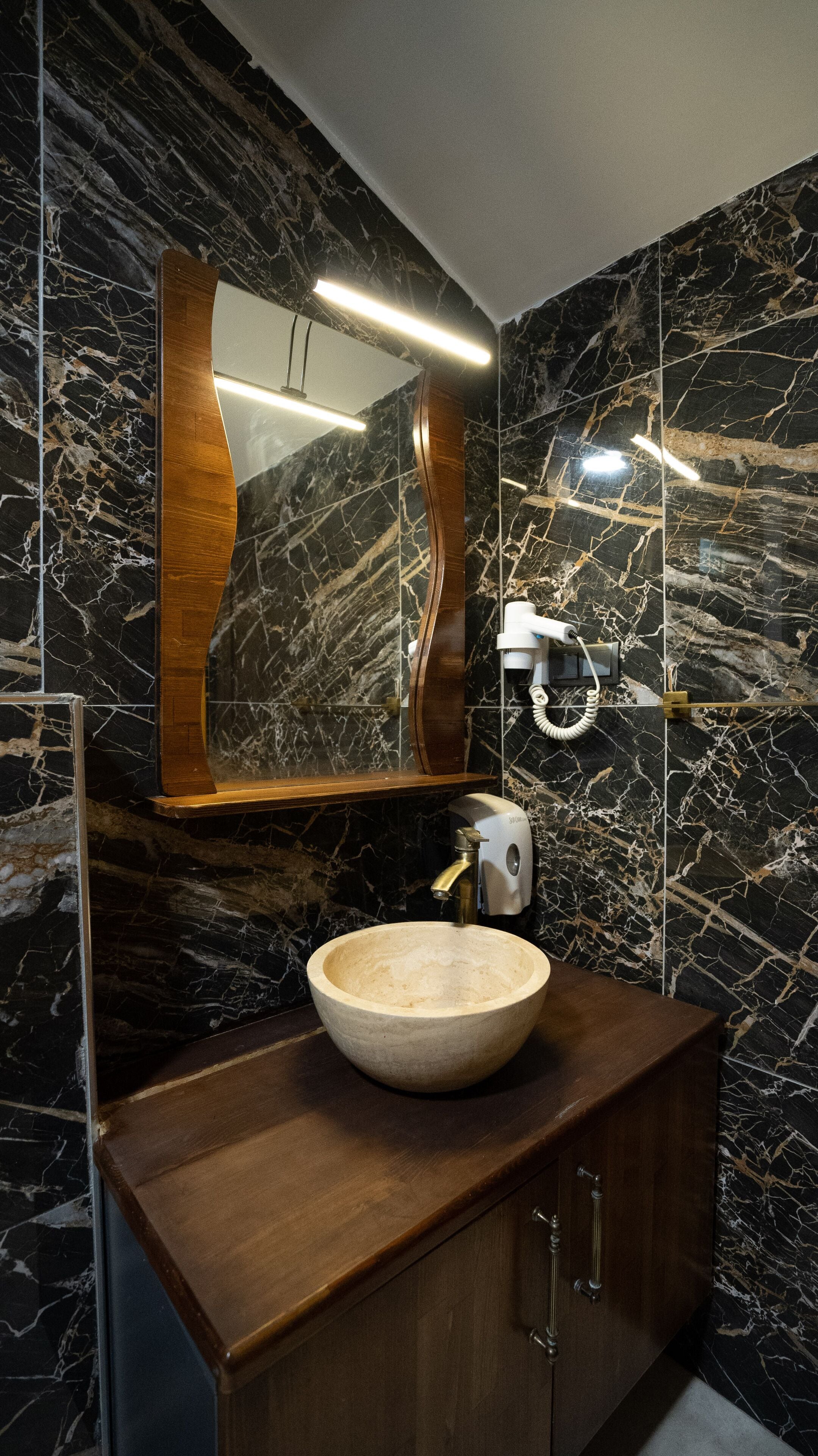 A bathroom with a marble wall, a wooden vanity, a large mirror, and a hairdryer.