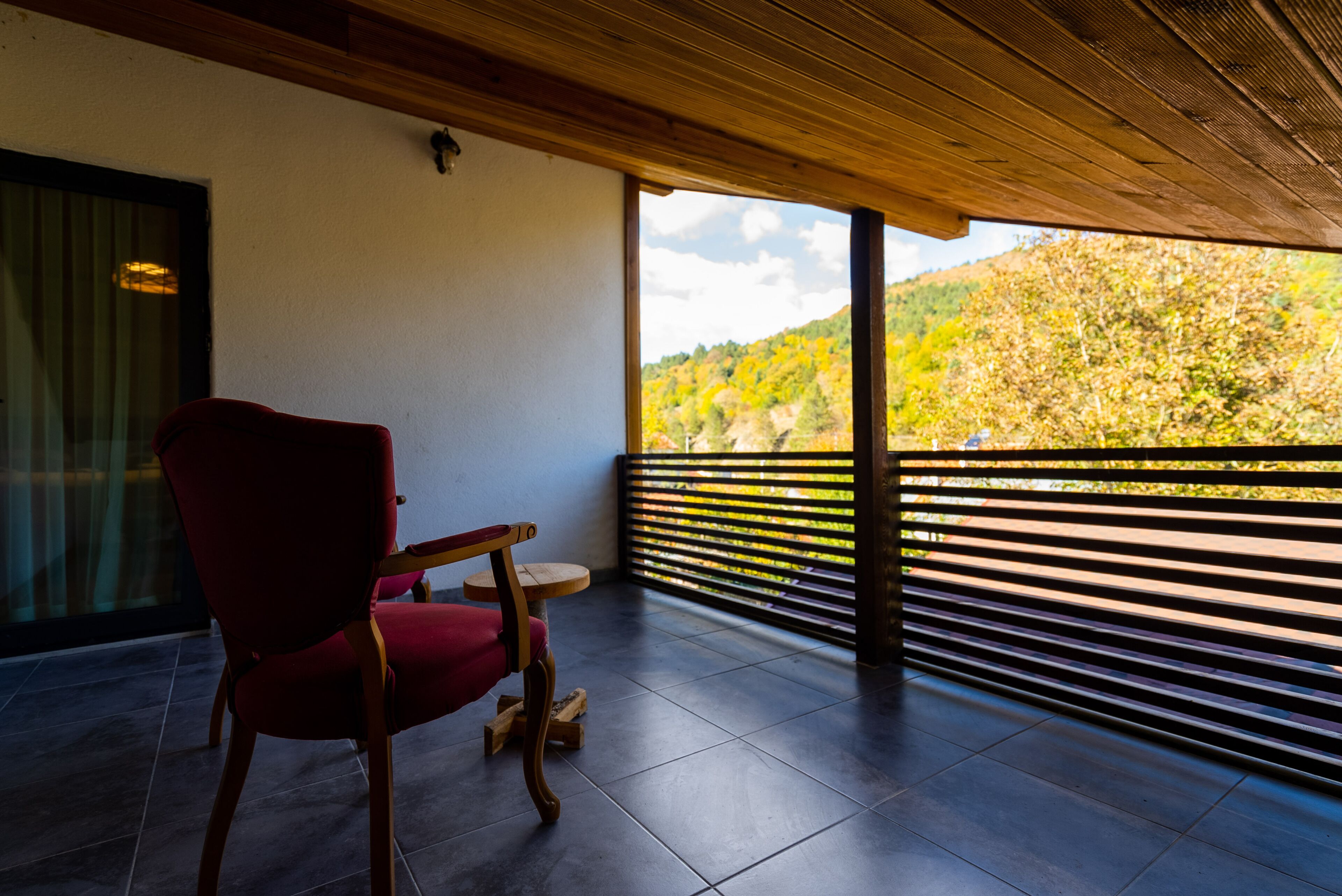 A balcony with a red chair, a wooden table, and a view of a hillside.