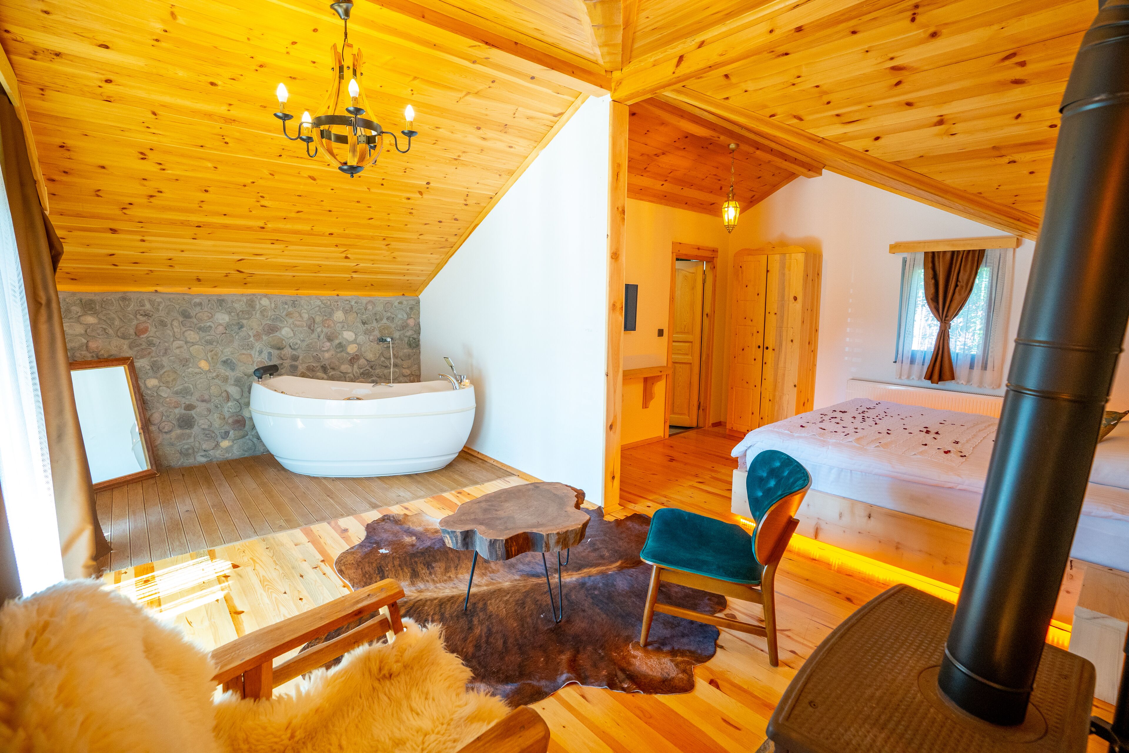 A wooden cabin interior with a stone wall, a large bathtub, a wooden table, a chair, and a bed.