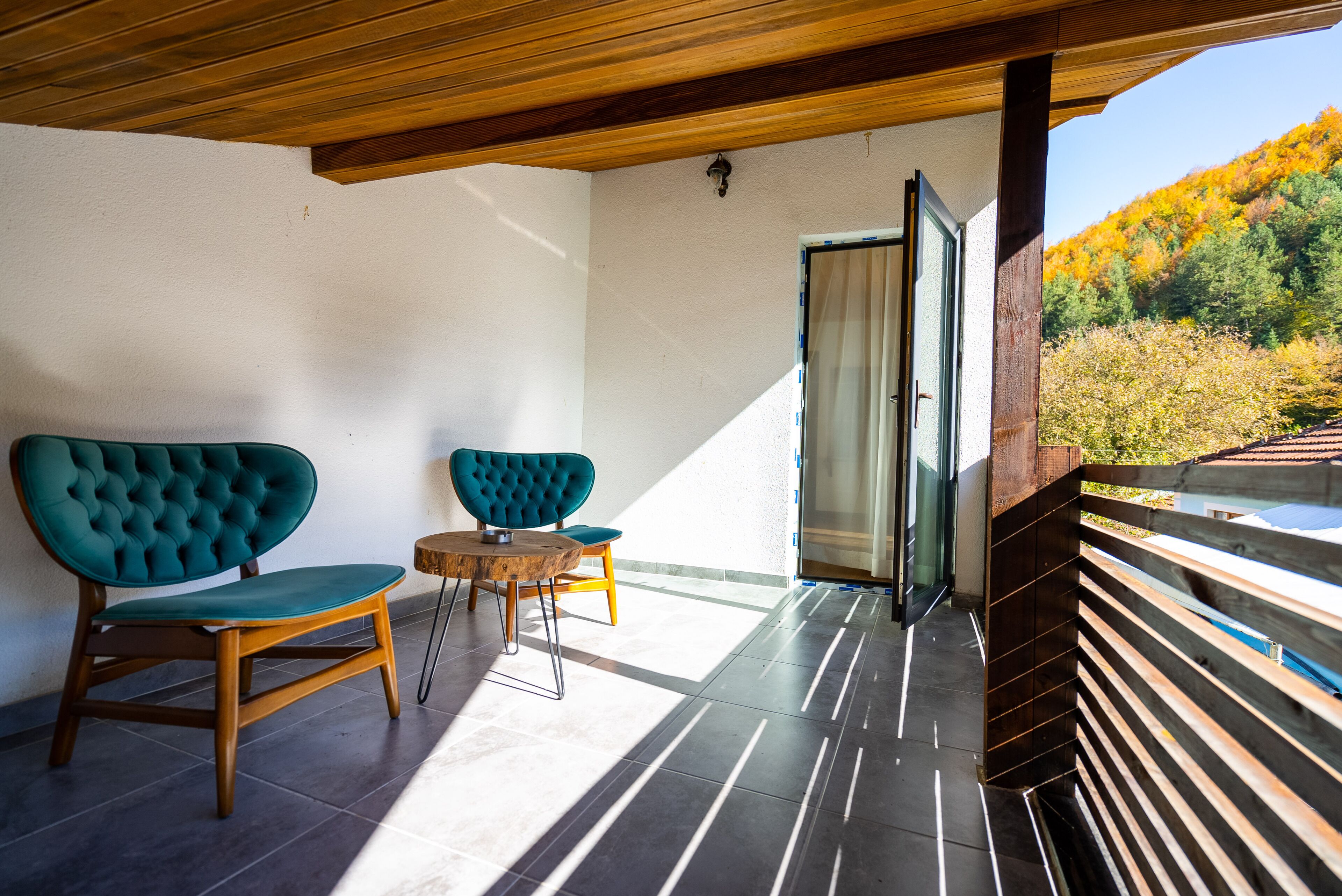 A balcony with two teal upholstered chairs and a small wooden table, a wooden ceiling, and a view of a forested hillside.