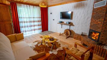 Uma sala de estar acolhedora com lareira, móveis de madeira, uma televisão e uma grande janela com cortinas.