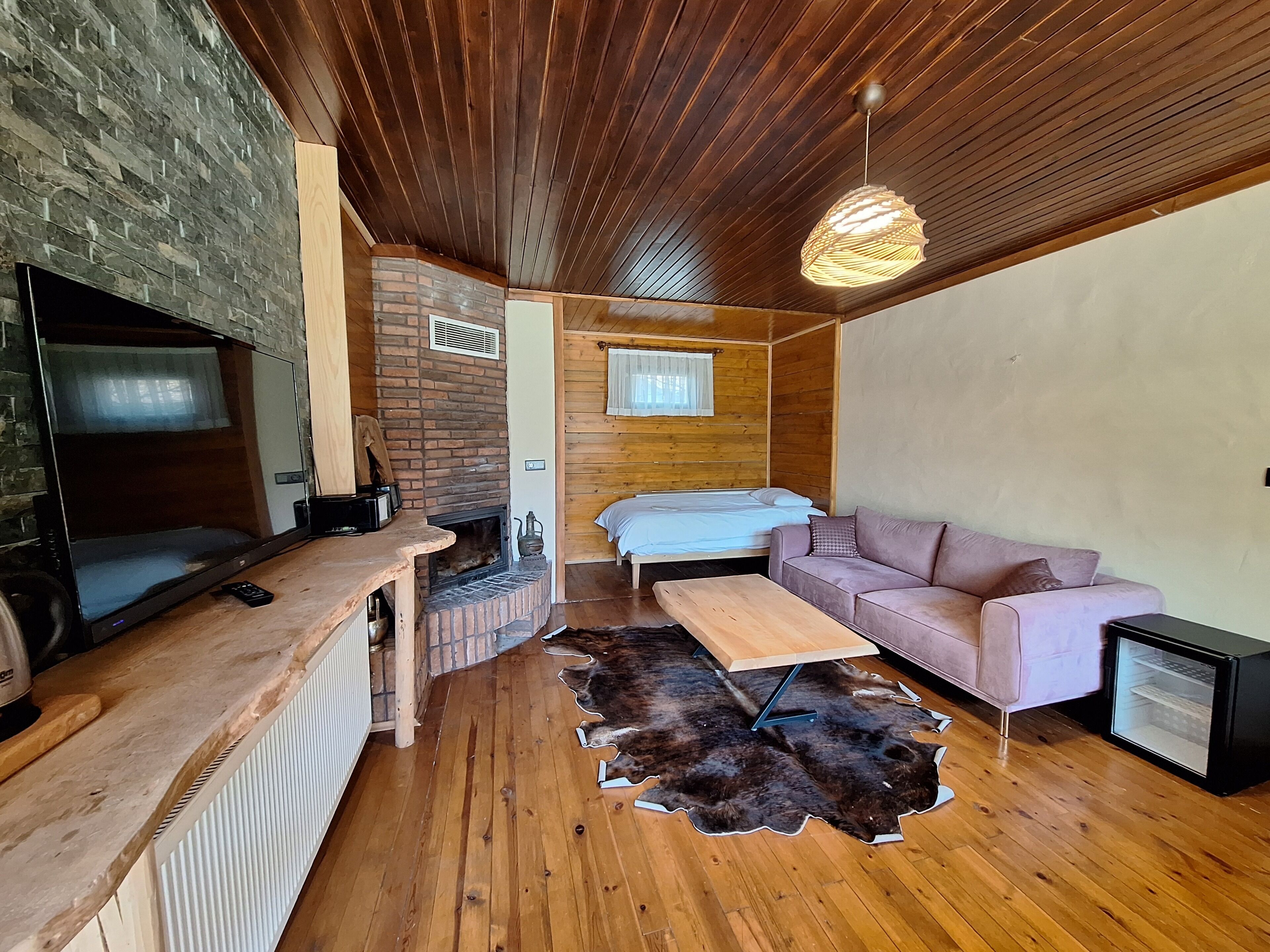 Ein gemütliches Blockhaus-Interieur mit Kamin aus Ziegelsteinen, Holzboden und einer kleinen Sitzecke inklusive Sofa, Tisch und Kühlschrank.