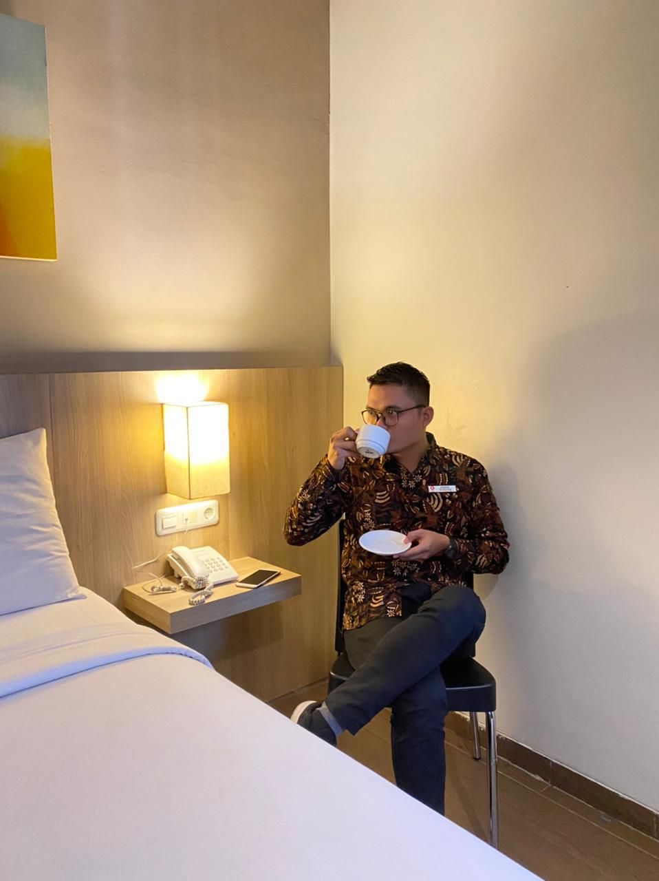 A hotel room with a bed, bedside table with a phone and lamp, and a person sitting on a chair drinking from a cup.