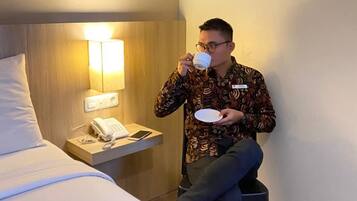 A hotel room with a bed, bedside table with a phone and lamp, and a person sitting on a chair drinking from a cup.