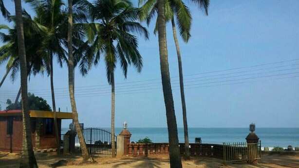 A coastal area with palm trees, a fenced gate, and a building with a red roof. - Hotel Killa Niwas (Malvan)
