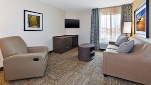 A hotel room with a beige sofa, a brown armchair, a flat-screen TV, a small round table, and a patterned carpet. - Candlewood Suites Louisville - NE Downtown Area by IHG (Louisville)