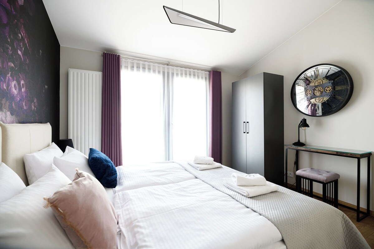 A modern hotel room with a large bed, a desk, a clock, and a window with curtains.