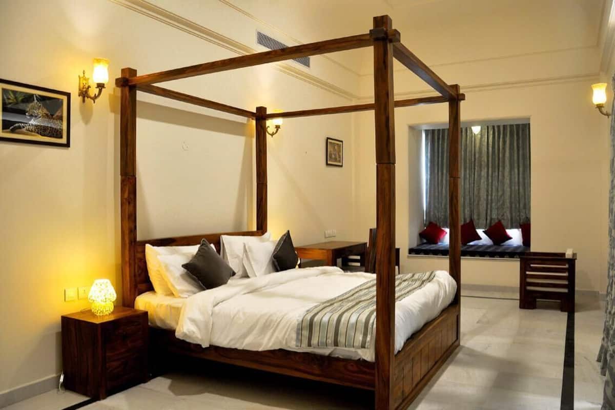 A four-poster bed with a wooden frame, a bedside table with a lamp, and a view of the room through a window with curtains.