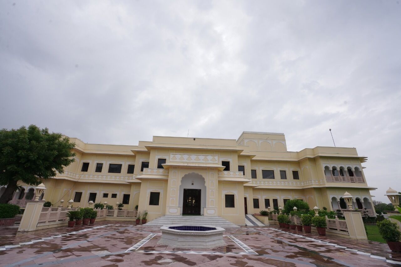 A large, multi-story building with a central entrance, surrounded by a courtyard with a fountain and potted plants.