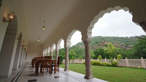 A covered outdoor seating area with a view of a green landscape and hills. - Hotel Raj Bagh Palace (Amer)