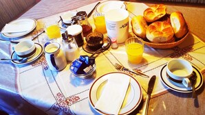 Une table de petit-déjeuner dressée avec des assiettes, des tasses, un panier de pain, une cafetière et une bouteille de jus de fruits.