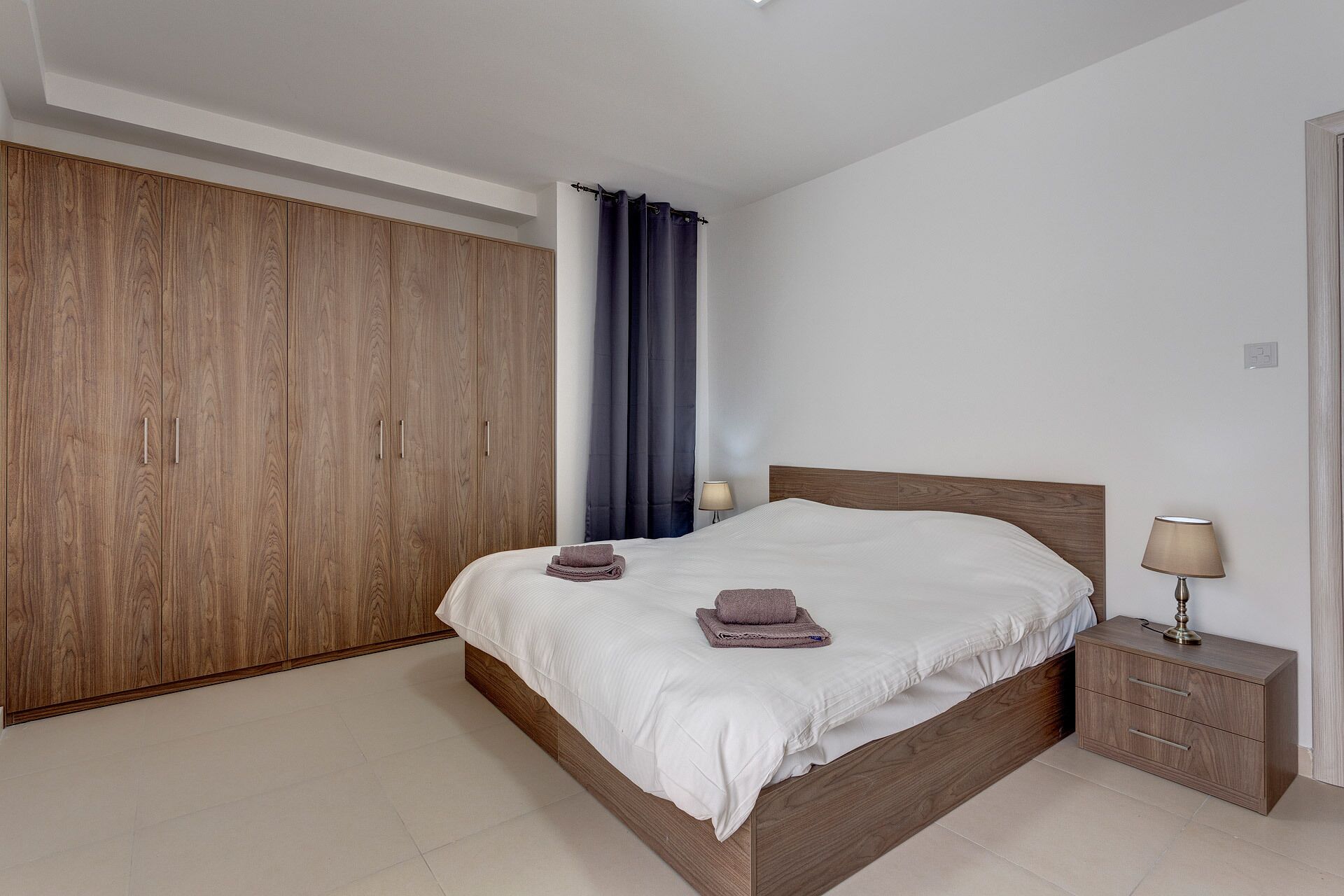 A bedroom with a wooden wardrobe, a bed with white bedding, a nightstand with a lamp, and a blue curtain.
