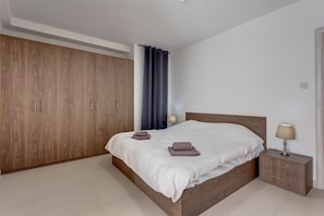 A bedroom with a wooden wardrobe, a bed with white bedding, a nightstand with a lamp, and a blue curtain. - Idyllic 1 Bedroom Apartment in Malta (Msida)