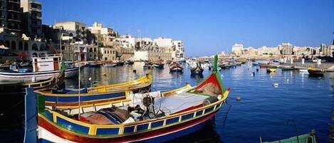 A harbor with boats docked, buildings in the background, and calm water.