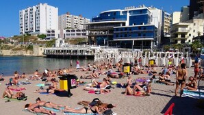 Una playa concurrida, gente tomando el sol y nadando, un muelle con barcos, y edificios al fondo.