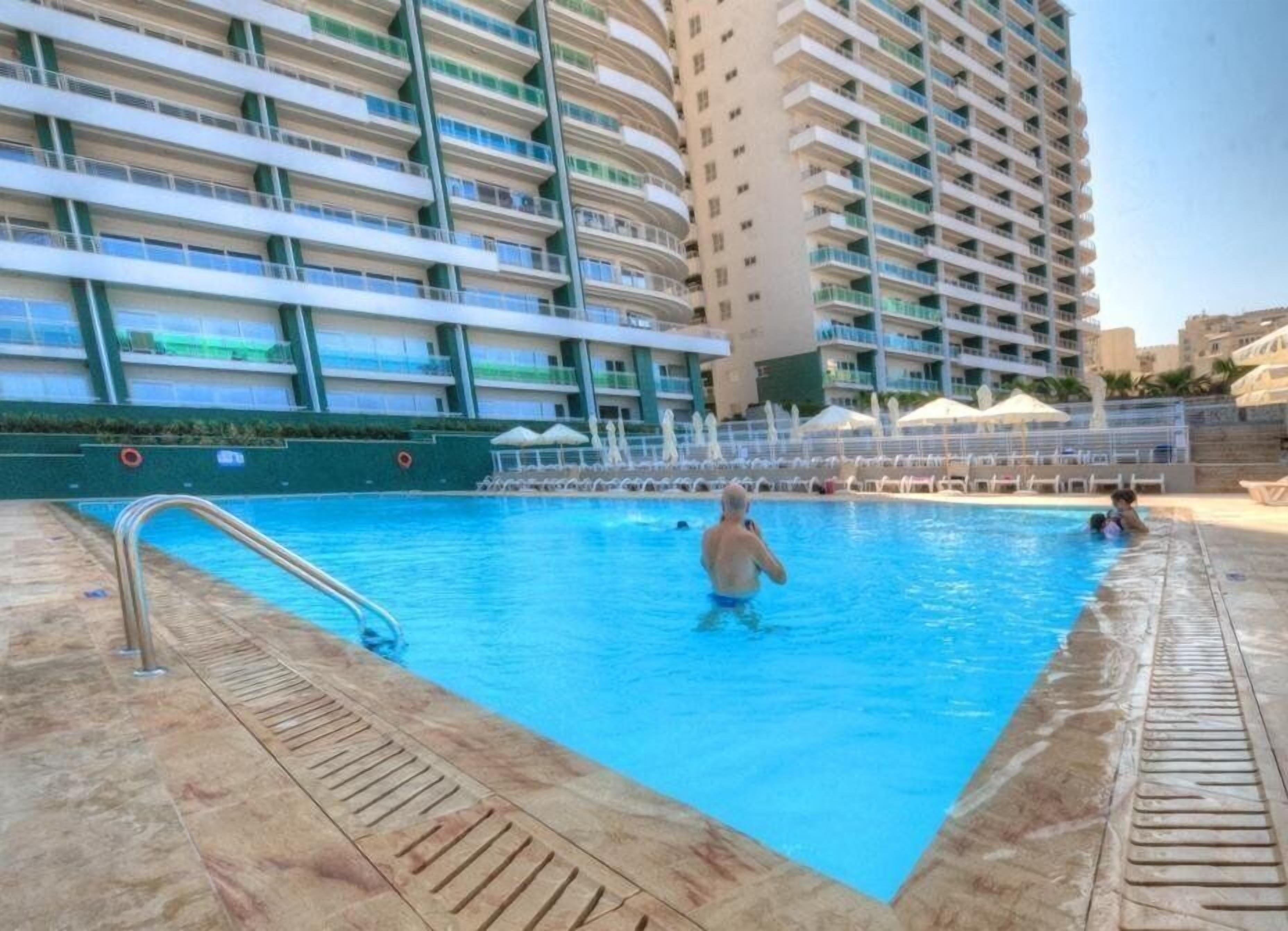 En swimmingpool med en metallstege, omgiven av en terrass med kakelplattor.