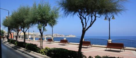 A coastal promenade with benches and street lamps, overlooking the sea and cityscape.