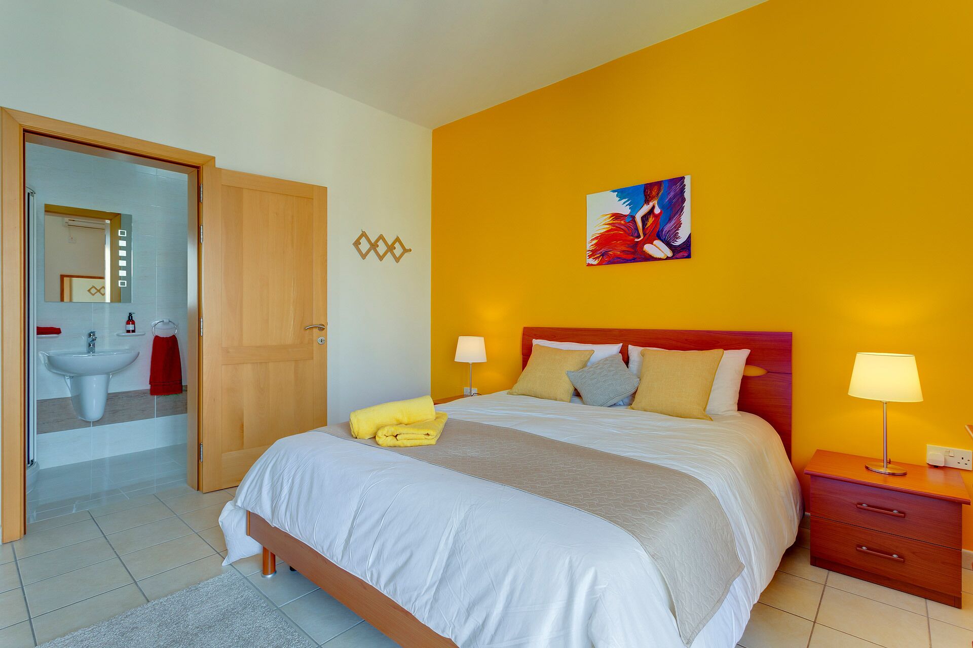 A hotel room with a bed, bedside tables, a nightstand, a mirror, a sink, and a towel rack.