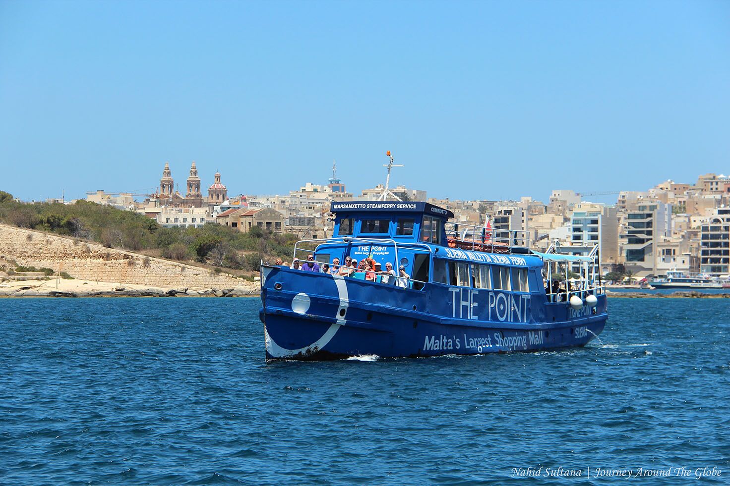 Eine blaue Fähre namens "The Point" mit der Aufschrift "Malta's Largest Shopping Mall" auf dem Rumpf, fährt auf dem Wasser.