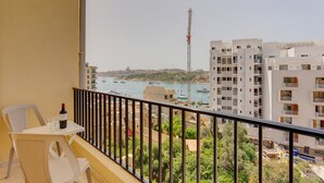Ein Balkon mit Tisch, Stühlen und einer Weinflasche. Blick auf einen Yachthafen mit Booten und einem hohen Turm in der Ferne.