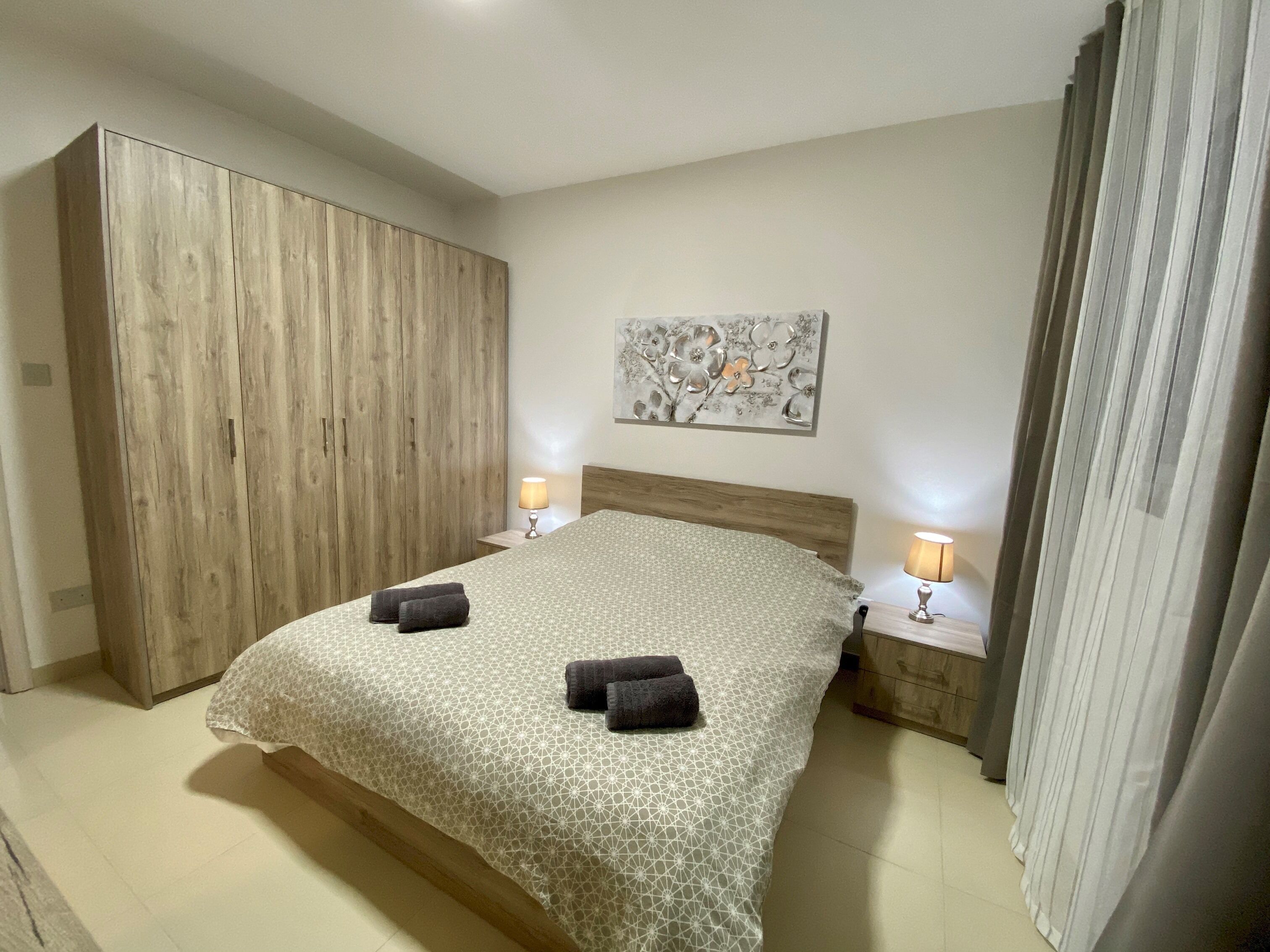 A bedroom with a large wardrobe, a bed with a patterned bedspread, two bedside lamps, and a floral artwork on the wall.