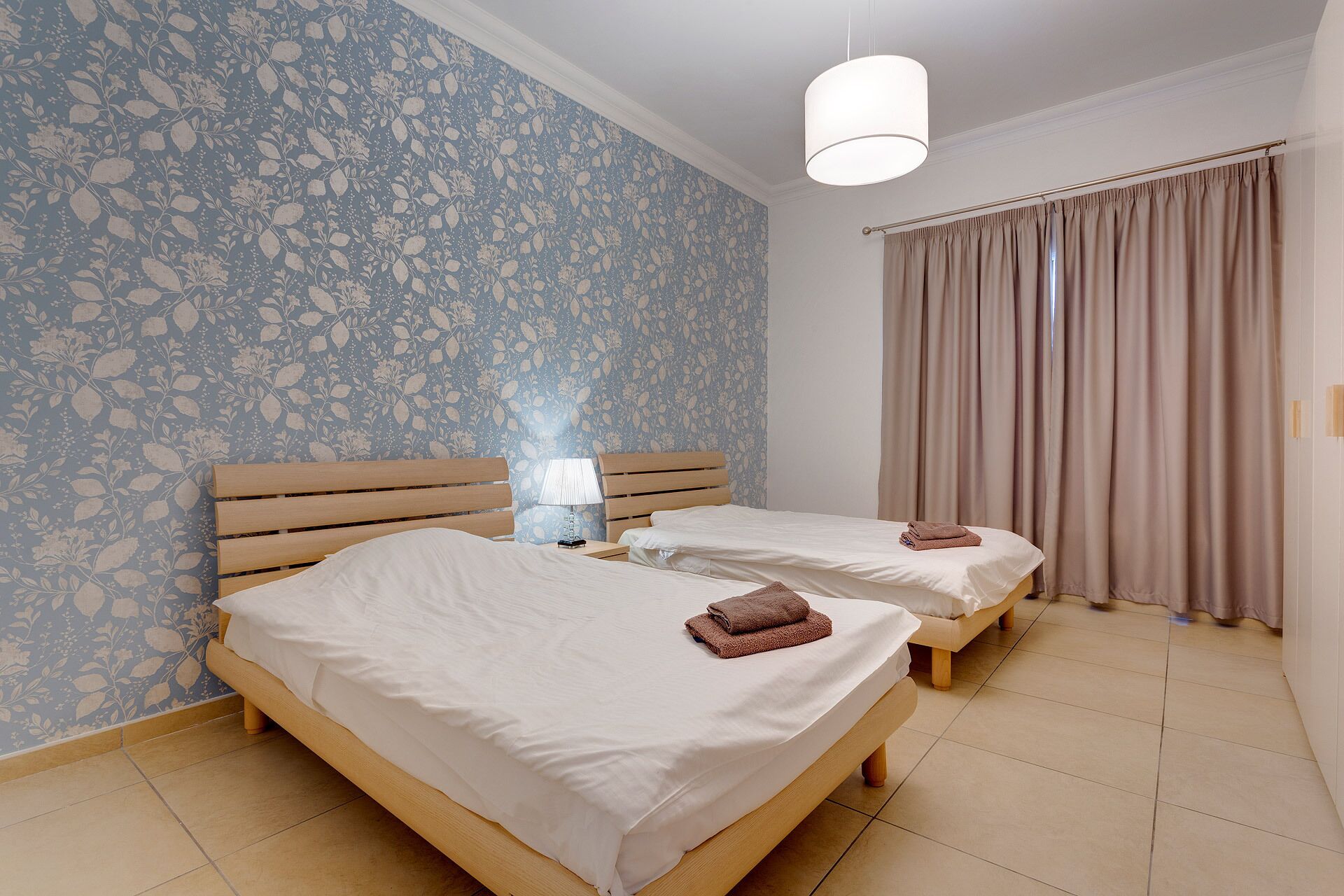 A hotel room with two wooden beds, a nightstand with a lamp, a window with curtains, and a floral wallpaper.
