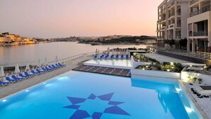 A rooftop pool with a geometric star design, surrounded by blue lounge chairs and umbrellas, overlooking a body of water and historic buildings. - Modern-luxury in Tigne Point w Pool, Best Location (Sliema)