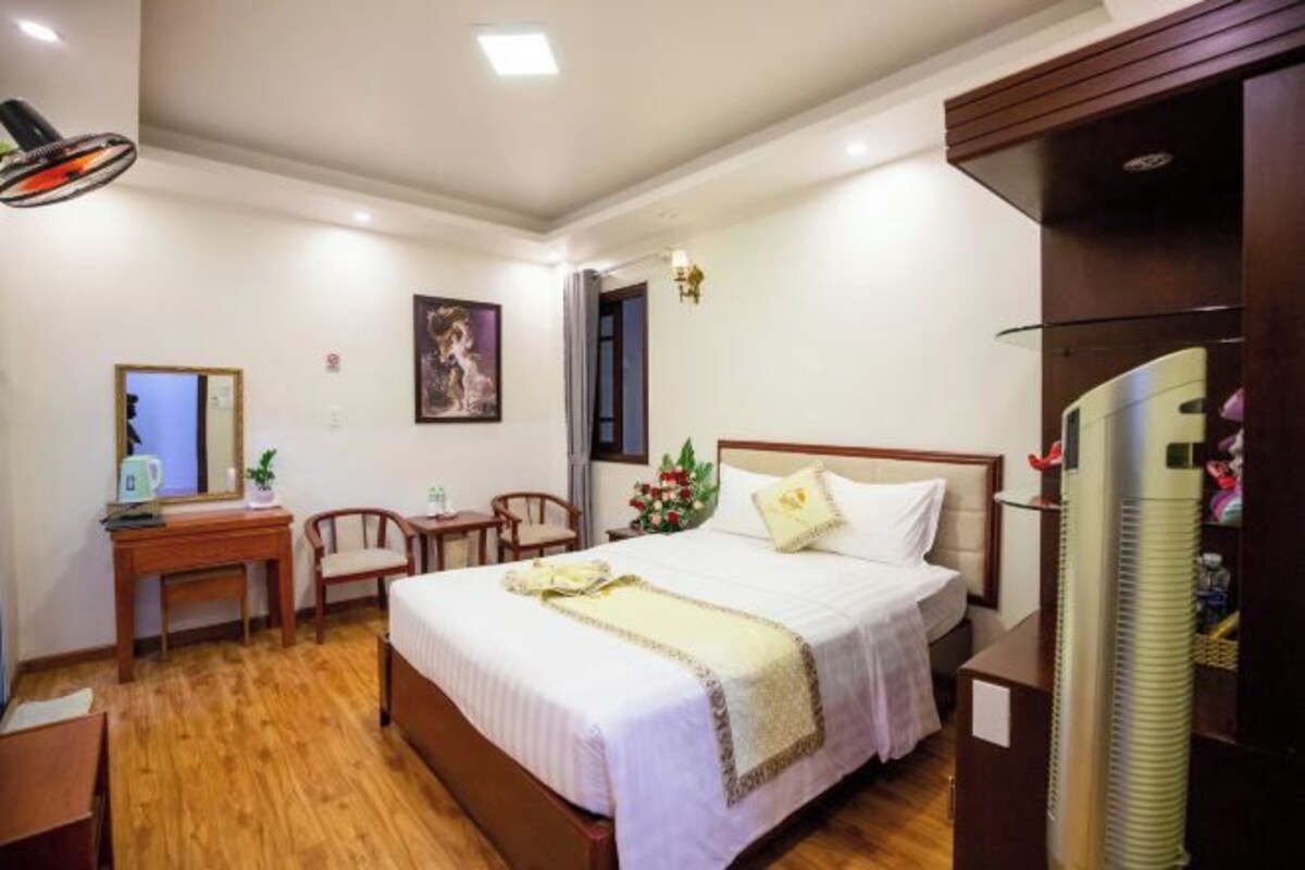 A hotel room with a bed, wooden desk, chair, and a small table with a lamp.
