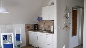 A compact kitchen with white cabinets, a coffee maker, and a sink. There is a glass dining table with blue chairs.