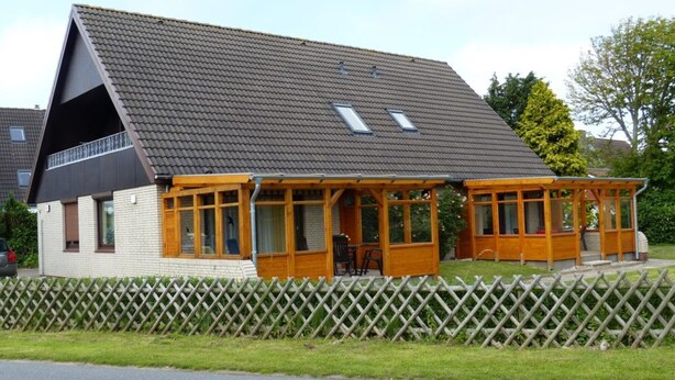 A two-story house with a sloped roof, a covered porch, and a fenced yard. - Holiday house with large garden, covered terrace, near the beach,  (Westerdeichstrich)
