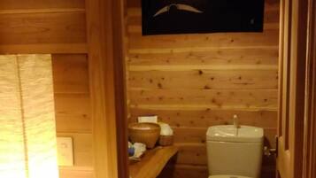 A wooden cabin interior with a wooden floor, a white toilet, a shelf with toiletries, and a hanging light fixture.