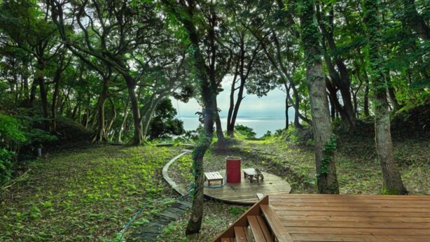 Une terrasse en bois avec un banc, une petite table et un tonneau rouge, entourée d’une végétation luxuriante et d’arbres, et offrant une vue sur une étendue d’eau.