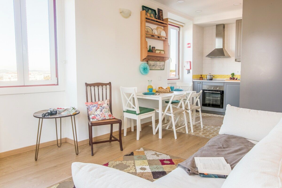 A cozy living room with a sofa, a small table, and a dining area with chairs and a kitchen in the background.