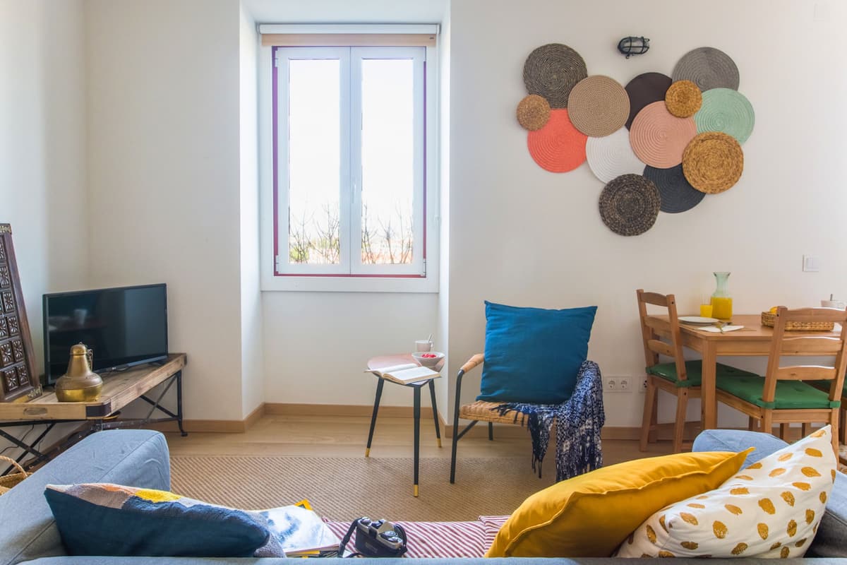 Una sala de estar con un sofá, una mesa de centro, un televisor, una mesa de comedor con sillas, una ventana y un adorno en la pared.