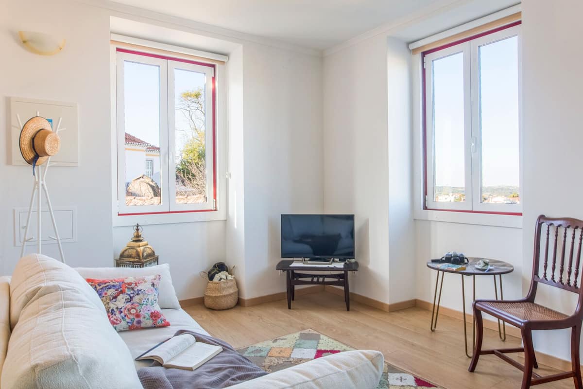 Una sala de estar acogedora con un sofá, un televisor, una mesita con una silla y dos ventanas con marcos rojos.