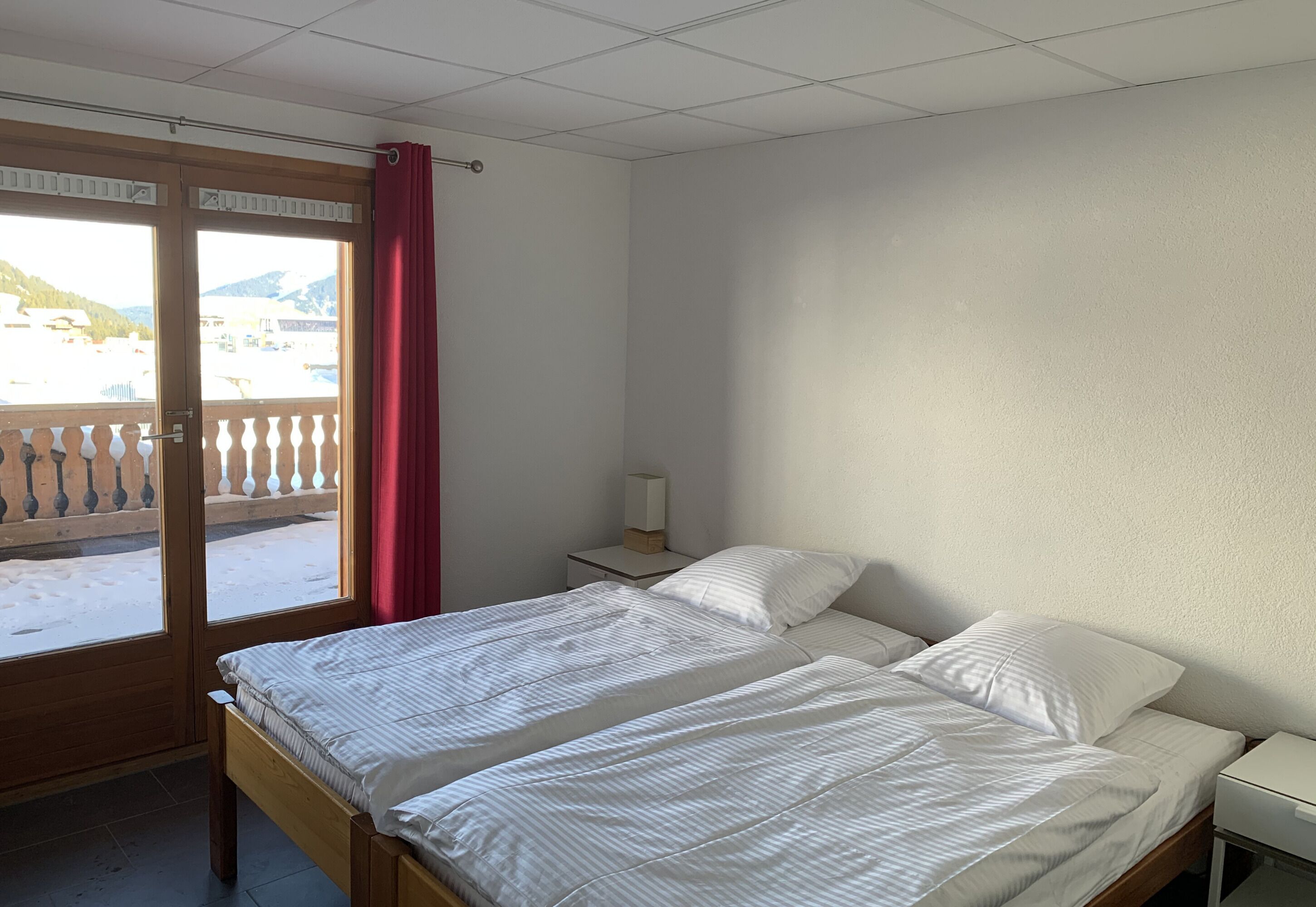 Habitación con dos camas, una ventana que da a un paisaje nevado, suelo de madera y una mesita de noche con lámpara.