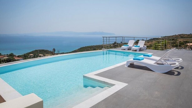 A rooftop infinity pool with clear blue water, white lounge chairs, and a view of the sea and distant hills. - Countrystyle Villa Bonora (Skala Kefalonias)