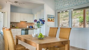 A dining area with a wooden table and wicker chairs, a kitchen in the background, and a window with a view of the outdoors. - Countrystyle Villa Bonora (Skala Kefalonias)