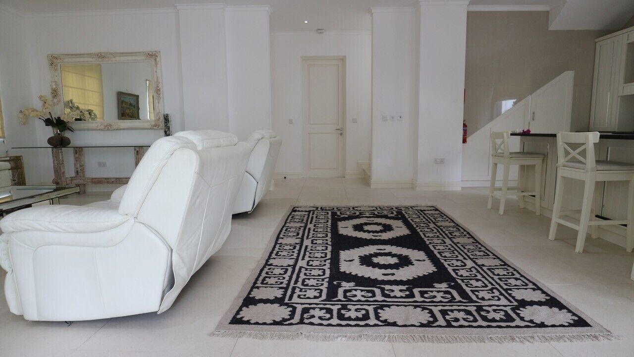 A spacious living room with a large white sofa, a patterned rug, and a dining area with white chairs.