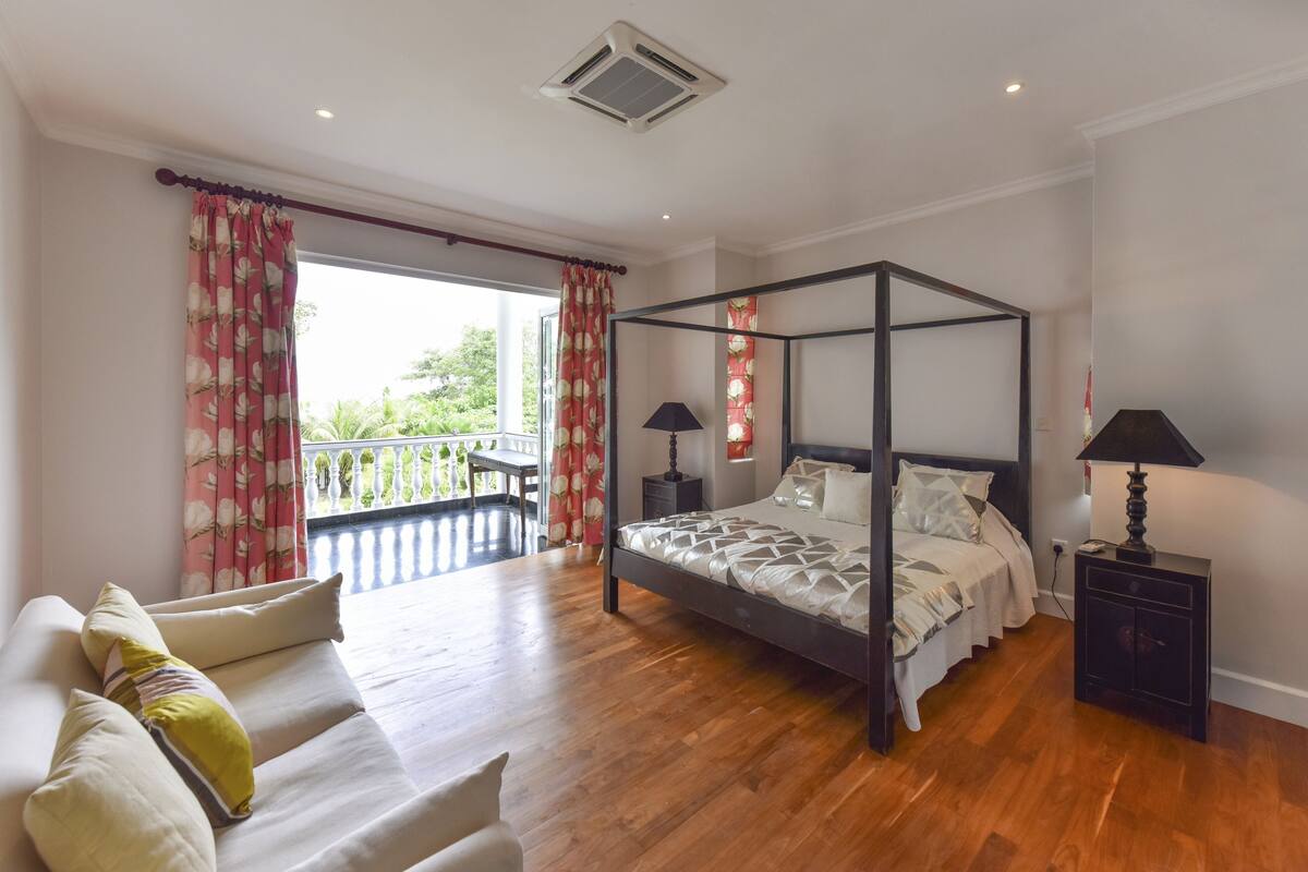 A four-poster bed with a canopy, a sofa with cushions, a small bedside table with a lamp, and a view of greenery through the window.