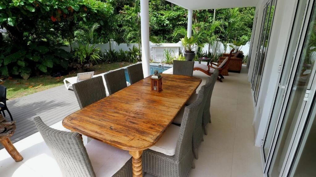 An outdoor patio with a wooden table and wicker chairs, surrounded by lush greenery and a pool.