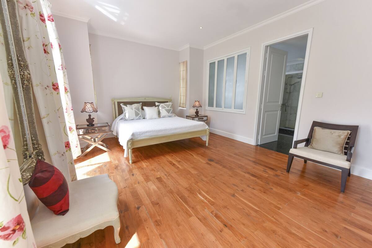 A bedroom with a wooden floor, a bed with white bedding, a bedside table with lamps, a chair, and a door leading to a bathroom.