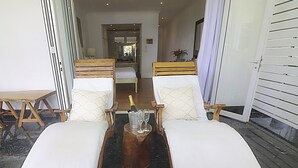Une pièce comprenant deux fauteuils en bois avec des coussins blancs, une petite table en bois sur laquelle se trouve une bouteille de champagne et des verres, et un lit en arrière-plan.