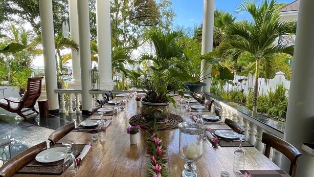 Une longue table à manger, dressée pour un repas, est agrémentée d’une guirlande décorative et entourée de chaises. La pièce offre une vue sur des plantes tropicales et est ornée d’une colonne blanche.