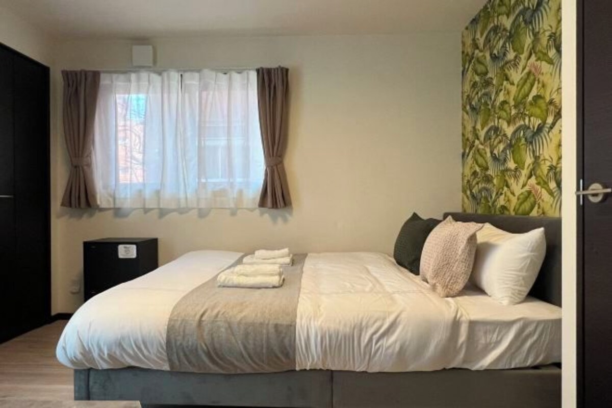 A neatly made bed with a blanket and pillows, a bedside table, and a window with curtains.